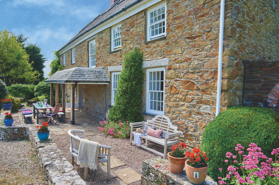 Sunny stone cottage patio with benches, table, potted flowers, and manicured shrubs under a blue sky.