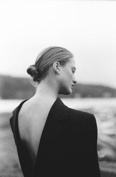 Black-and-white portrait of a woman with a bun and bare back in a dark jacket, looking away by a blurred waterside landscape.