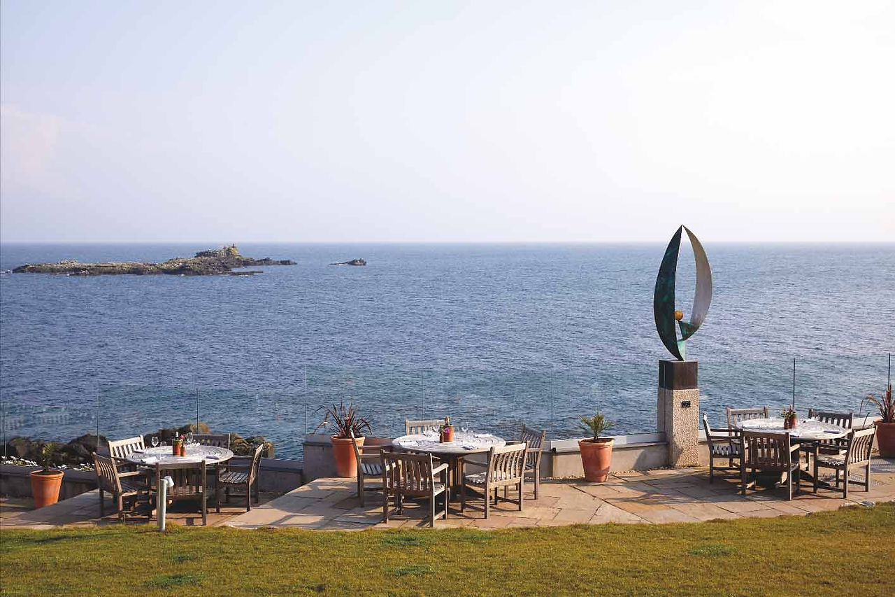 Seaside patio with empty tables and chairs, potted plants, and a modern sculpture overlooking calm ocean and a rocky island.