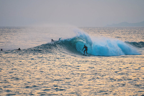 Surfer rides inside a blue curling wave at sunset, with other surfers waiting nearby on a calm ocean.