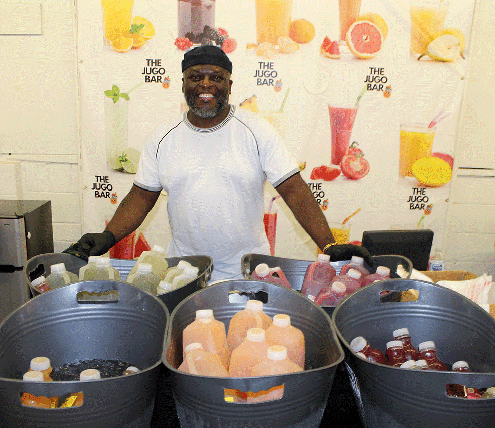 William with plastic baskets of juice quarts in front of him. Behind him is a backdrop with pictures of smoothies and the words "The Jugo Bar"