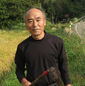 田んぼ脇のあぜ道でインタビューを受ける小白倉区長の田中一男さん