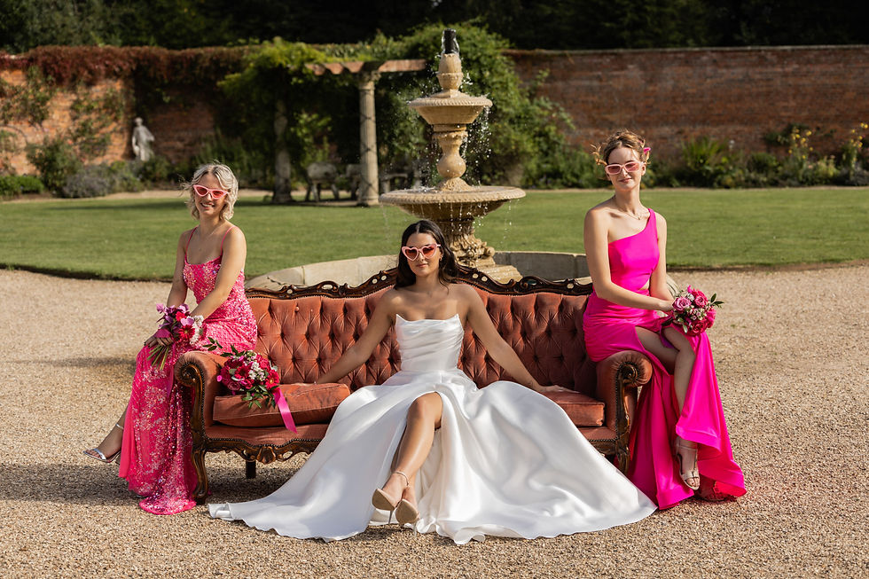 Stylish Fine art wedding photo of bride posing on large pink sofa, with bridesmaids in pink dresses all wearing pink heart sunglasses in front of water fountain at Elmhay Park Somerset