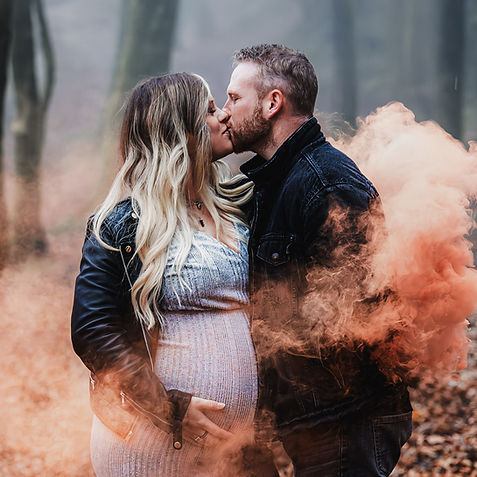 Spooky foggy woodland maternity photoshoot with fun orange smoke bombs, mother and father to-be sharing kiss