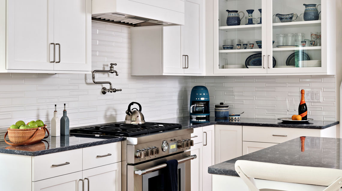 Spacious white coastal kitchen with a large island and tall bar-style chairs, featuring dark quartz countertops, a stainless steel refrigerator and gas range, and a farmhouse-style sink with a Kohler Artifacts faucet. Wood floors add warmth and character to the open design.