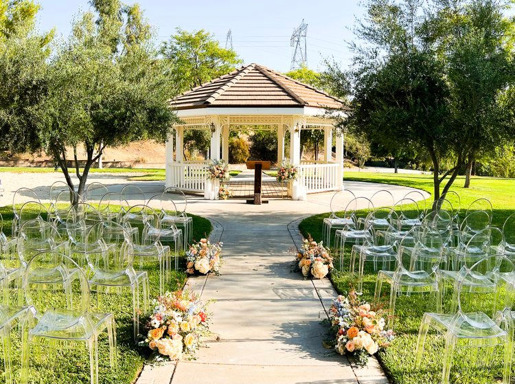 Sunny outdoor wedding setup with a gazebo in the background, clear chairs, and floral arrangements lining the aisle. Peaceful and elegant mood.