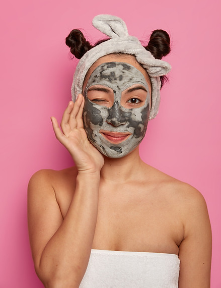 Studio shot of lovely young ethnic woman applies face mask with mud, winks eye, touches ch