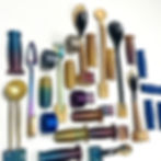 A variety of objects, likely 3D printed, are arranged on a white surface. The objects vary in color and shape. There are metal-handled utensils, including spoons and a fork, with colors ranging from gold and bronze to iridescent blues, purples, and greens. Several appear to have handles with a textured or patterned surface. Some items that resemble rolls of tape or coiled wire are also visible in different colors. Metal objects in brass-colored and dark bronze tones are also present. The items appear to be diverse tools or components. They are arranged in an overlapping, somewhat artistic, or perhaps functional layout.