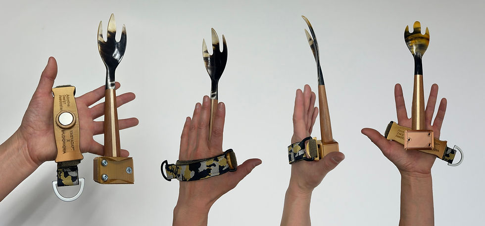 Four hands are holding a small, specialized utensil setup. The utensil consists of a fork-like tines on top of a long, slender handle, similar to a small, tapered knife. The handles appear to be made of a light brownish-gold colored material, possibly wood or a composite, with a black and gold pattern. The utensils are each affixed to a dark-colored wristband or strap with a camouflage-like pattern. A small metal ring and possibly a button or magnet is seen on the wristband portion. The hands are positioned in different angles, showcasing how the utensils might be held and used. The background is a plain, light gray or white.