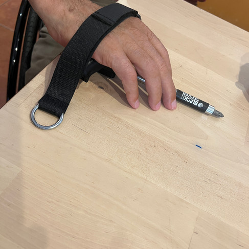 A person's hand is positioned on a light beige wooden table. The hand is wearing a black wrist support or splint with a metal ring attached.  A small black screwdriver or similar tool lies on the table near the hand. The person's forearm and upper arm are visible, and they're wearing a maroon colored shirt.  The lighting is natural and appears to be indoor.  The wooden surface shows wood grain.