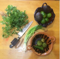 A light beige wooden surface is the backdrop for various ingredients arranged for a culinary preparation. A bunch of fresh cilantro is positioned at the upper left.  A small, dark-brown, handmade bowl, with a slightly rustic appearance and handles, contains several avocados and limes. Next to the bowl of avocados and limes, a small, round, terracotta-colored bowl with a unique, patterned design holds a few green tomatillos and a jalapeño pepper.  A bunch of green scallions/spring onions lies beside and underneath the terracotta bowl. A chef's knife with a dark handle is placed diagonally on the surface, between the cilantro and the other ingredients.  A thin, clear piece of string is tied around the scallions.