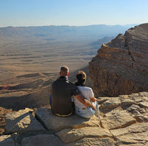זוג יושב על קצה מצוק וצופה בשקיעה במכתש רמון בטיול רייזר רמון