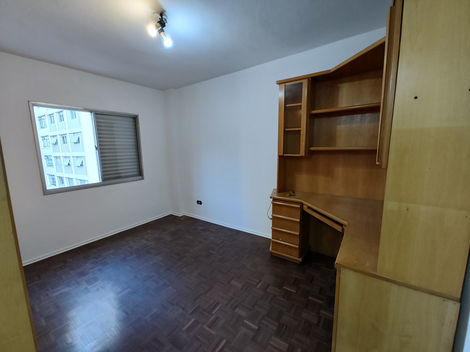 Sala de estar vazia com piso de madeira, paredes brancas e grande janela iluminando o ambiente.