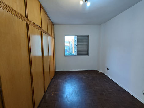 Quarto vazio com piso de madeira, paredes brancas e janela deixando entrar luz natural.