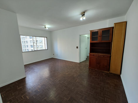 Sala de estar vazia com piso de madeira, paredes brancas e grande janela iluminando o ambiente.