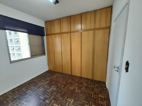 Quarto vazio com piso de madeira, paredes brancas e janela deixando entrar luz natural.