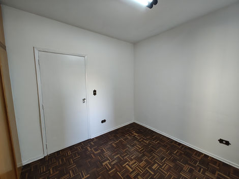 Quarto vazio com piso de madeira, paredes brancas e janela deixando entrar luz natural.
