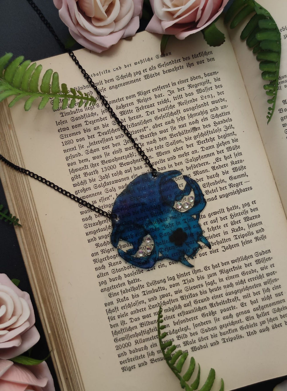 Blauer Totenkopf-Anhänger auf Buch mit Text, umgeben von Rosen.