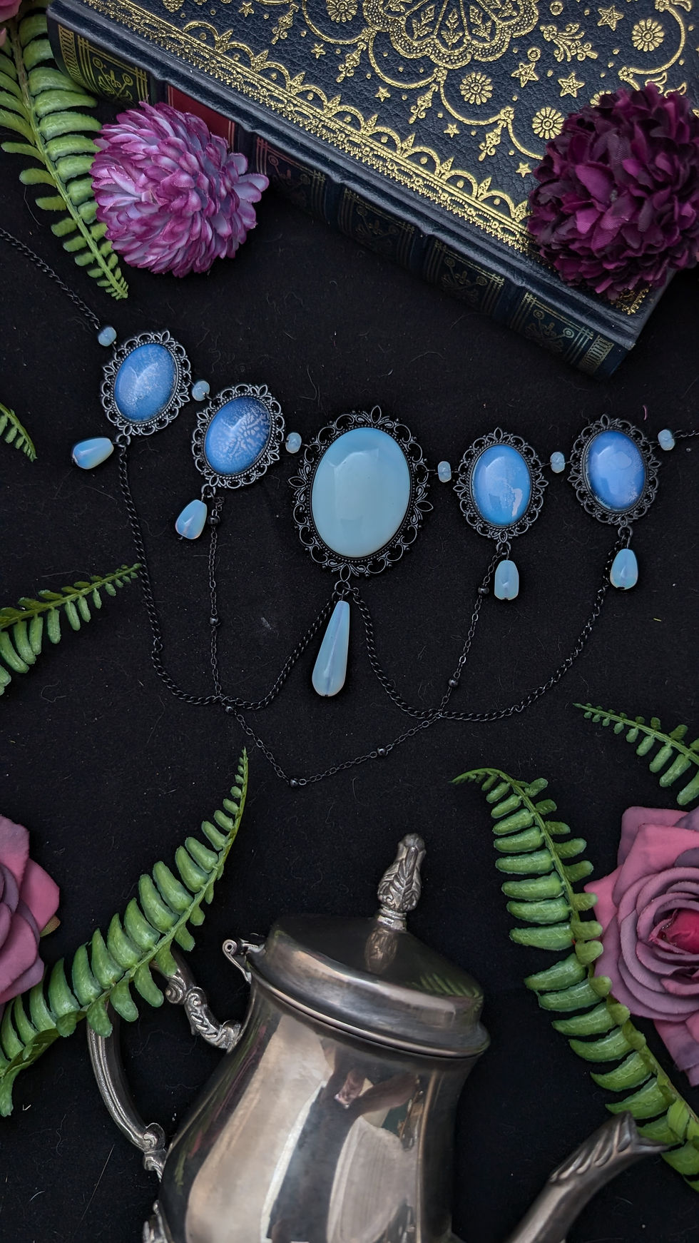 Blaue Edelstein-Halskette auf dunklem Stoff mit Blumen, Buch und Teekanne.
