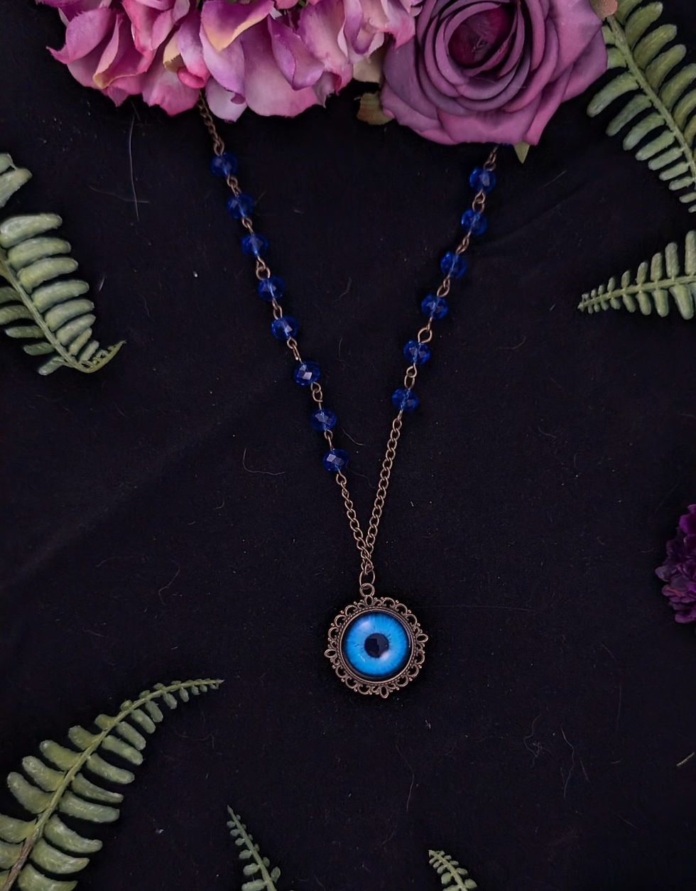 Blaue Halskette mit Augen-Anhänger, umgeben von lila Blumen und Farnen.