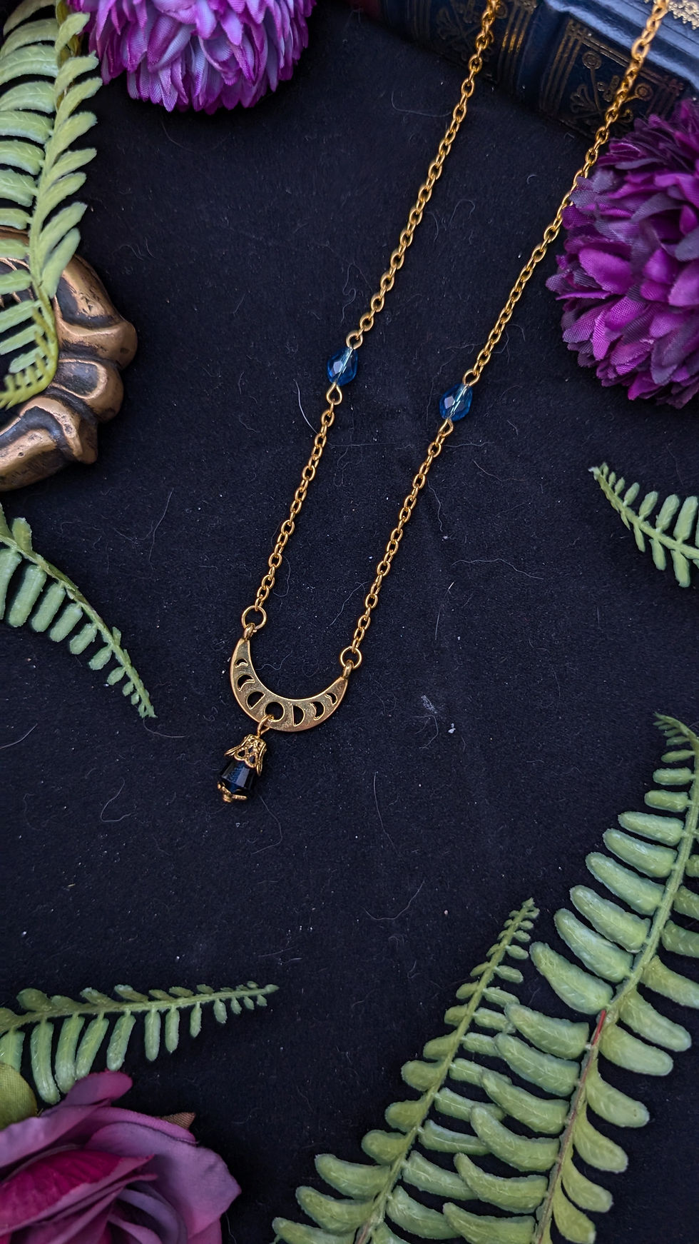 Goldkette mit Halbmond-Anhänger, blauen Perlen, umgeben von Farnen und Blumen.