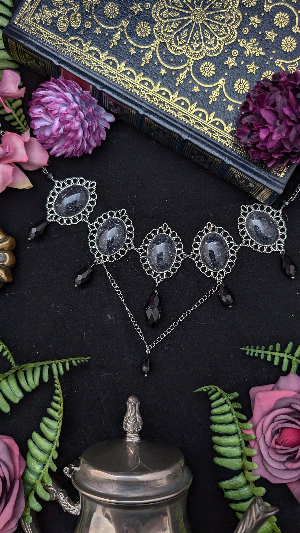 Gotische Halskette mit dunklen Steinen, umgeben von Buch und Blumen.