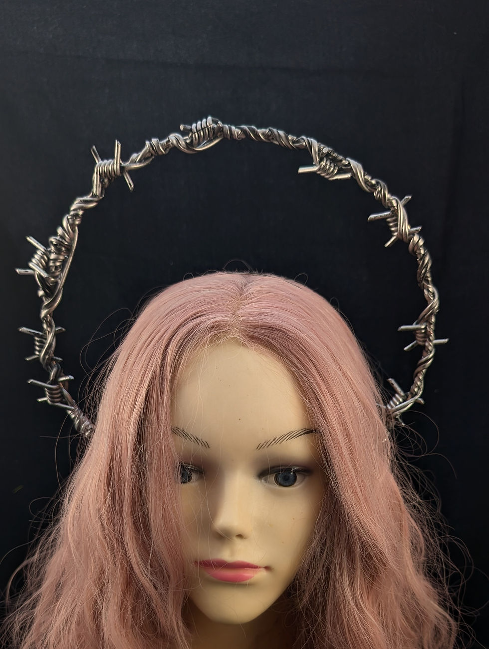 Mannequinkopf mit pinken Haaren trägt Stacheldrahtkrone vor schwarzem Hintergrund.