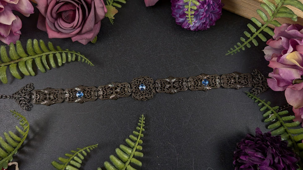 Dunkler, verzierter Halsreif mit blauen Edelsteinen, umgeben von Blumen und Farnen.