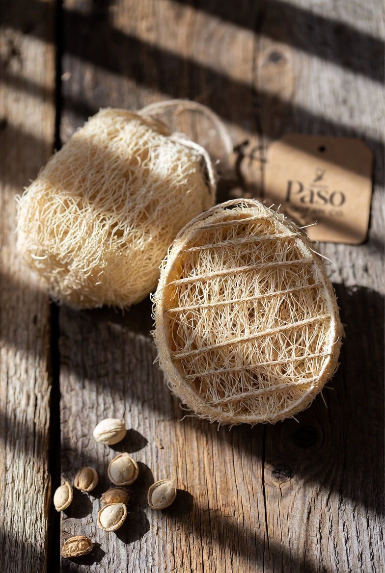 Close-up of whole and cut natural loofah sponges from Paso Loofah Co. showing fibrous interior texture on rustic wooden background with seeds