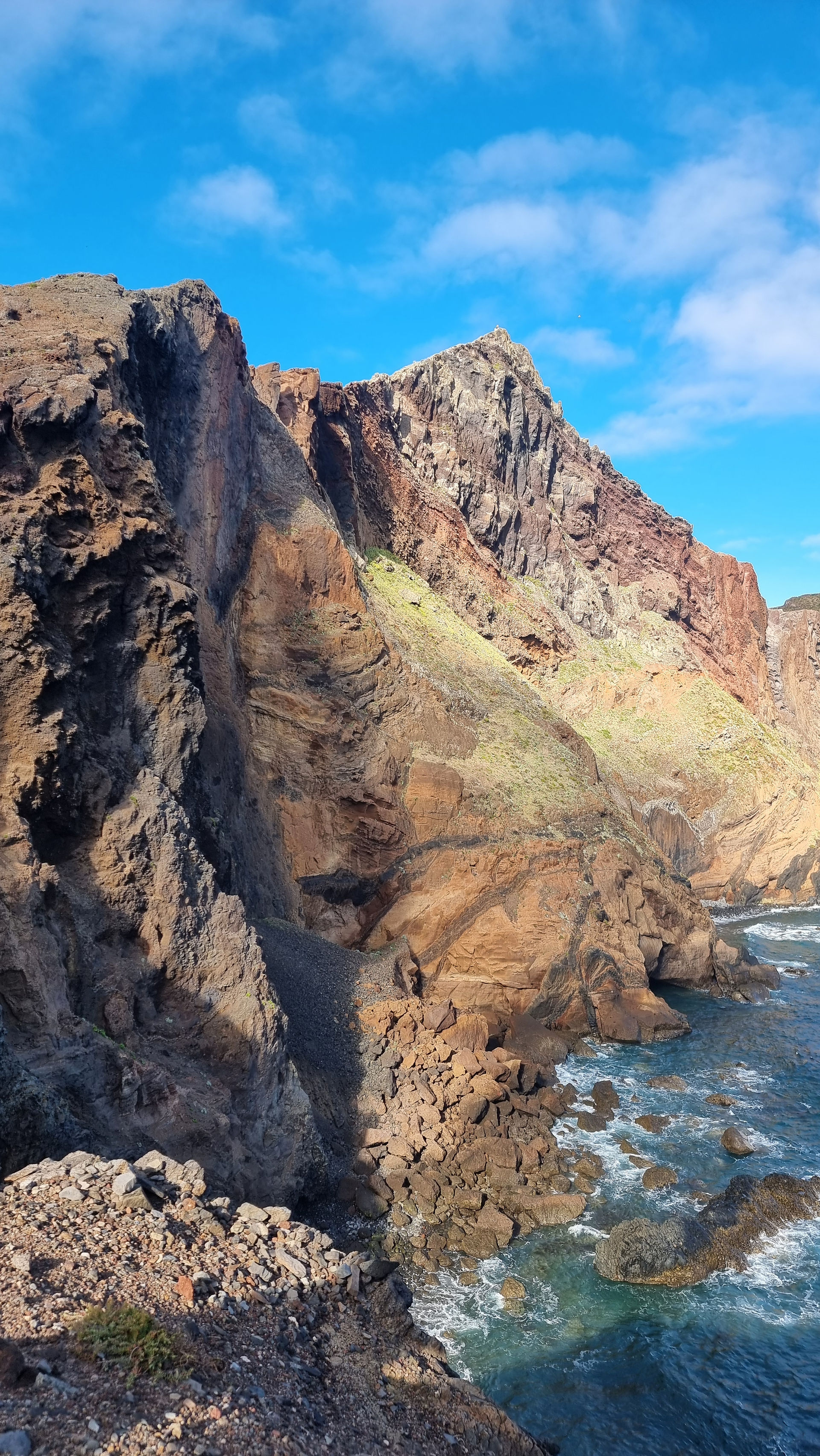 Steile vulkanische Felsklippen an der Küste Madeiras mit rötlich-braunen Gesteinsschichten, die direkt zum Atlantik abfallen, während Wellen gegen die Felsen schlagen und ein klarer blauer Himmel die Landschaft überragt.