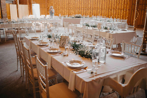 Svatební hostina v interiéru Stodoly v meruňkovém sadu s bílými chiavari židlemi, světelnou stěnou a prostřenými tabulemi s eukalyptem.