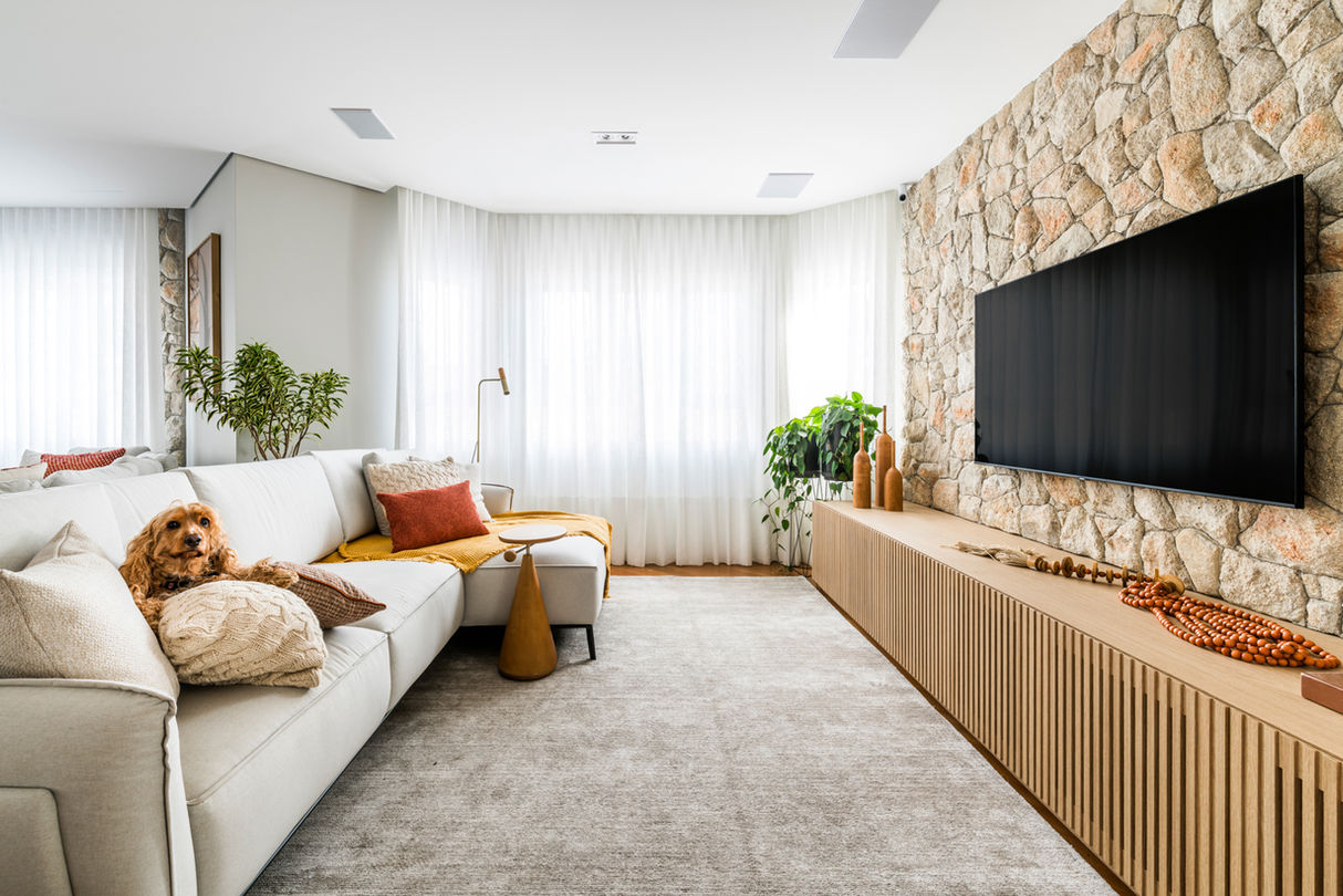 sala de tv em tons claro, com tv, rack em madeira, sofá bege, objetos em tons terracota e um cachorro sentado no sofá