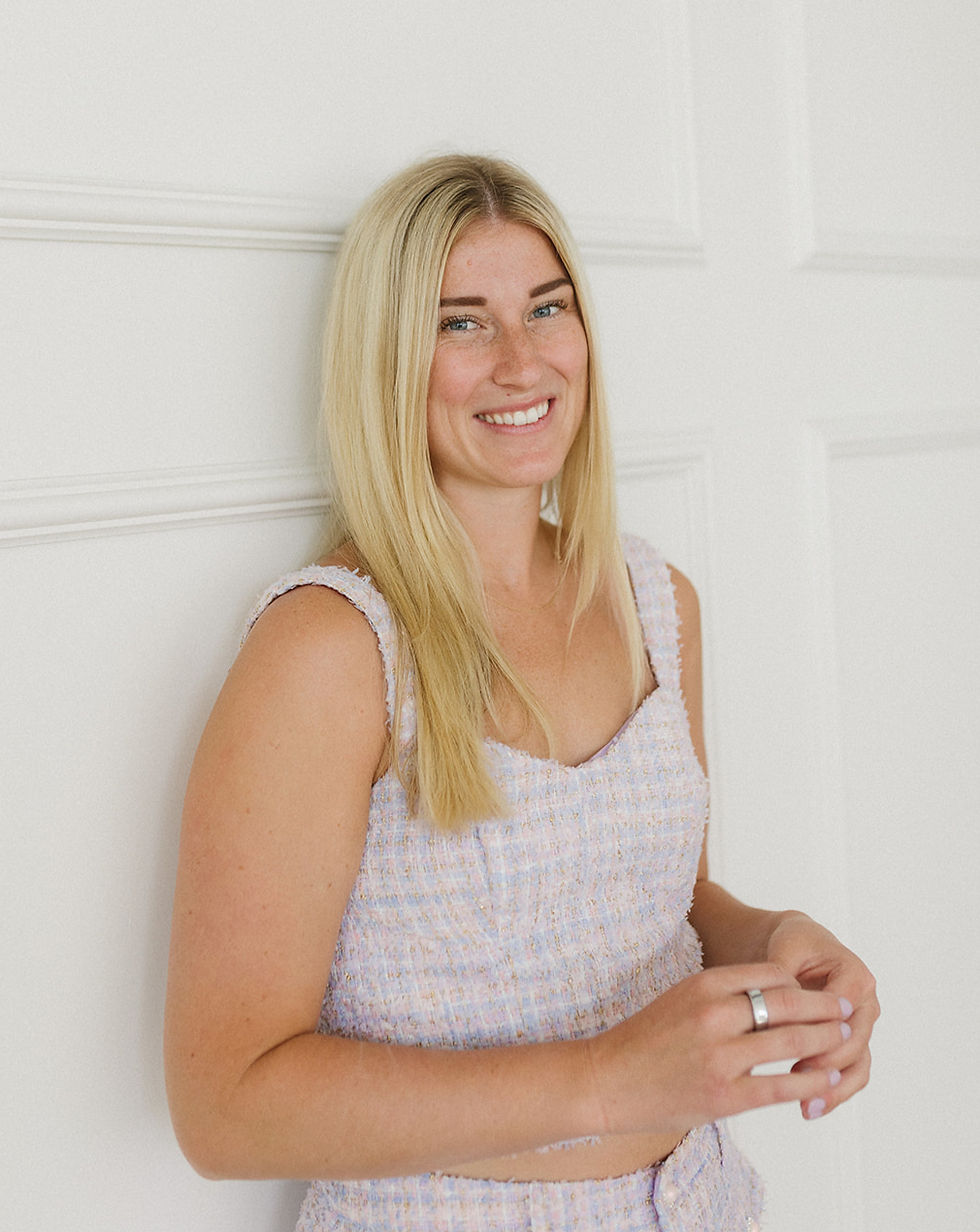 A white woman with blonde hair leans against a wall wearing purple.