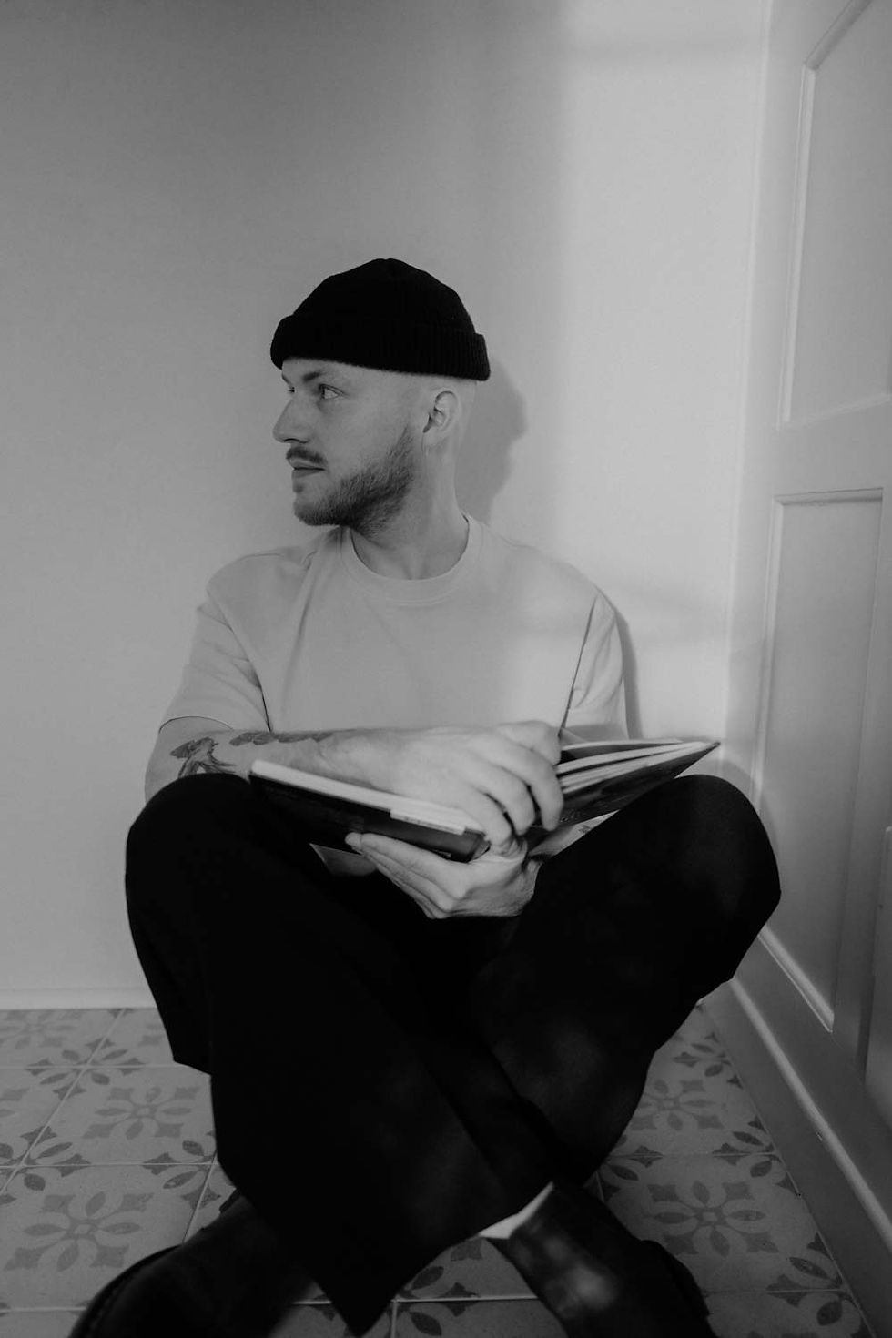 Porträt von Michael Christen, sitzend am Boden mit einem Buch in der Hand, seitlich blickend, in Schwarz-Weiss aufgenommen.