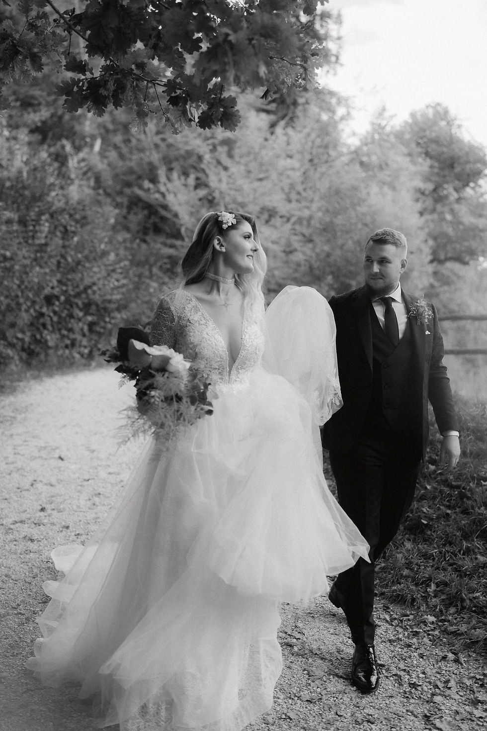 Die Braut läuft mit dem Brautkleid in der Hand neben dem Bräutigam und lacht ihn an. Aufgenommen in schwarz und weiss.