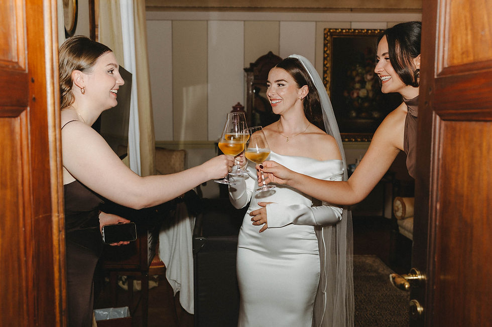 Drei Frauen stossen in eleganten Kleidern mit Wein an. Hintergrund zeigt ein Blumenbild und ein Sofa. Fröhliche Stimmung.
