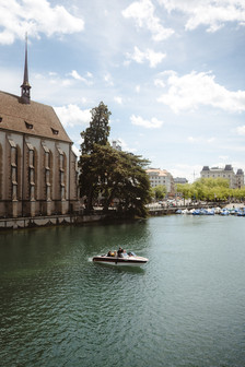 Kleines Boot auf der Limmat in Zürich, links eine Kirche mit spitzem Turm, im Hintergrund Boote am Ufer.