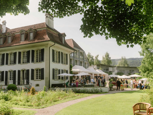 Schloss Hünigen mit rotem Dach, umgeben von Garten und Bäumen. Menschen feiern unter weißen Pavillons. Sommerliche, fröhliche Stimmung.