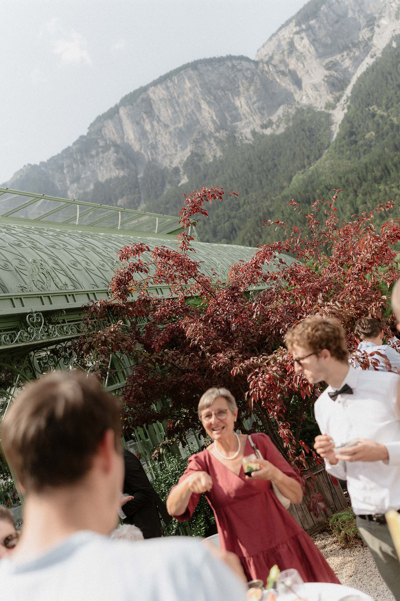 Lachende ältere Hochzeitsgästin im roten Kleid unterhält sich mit jungen Gästen vor dem Glaspavillon, im Hintergrund Berge.