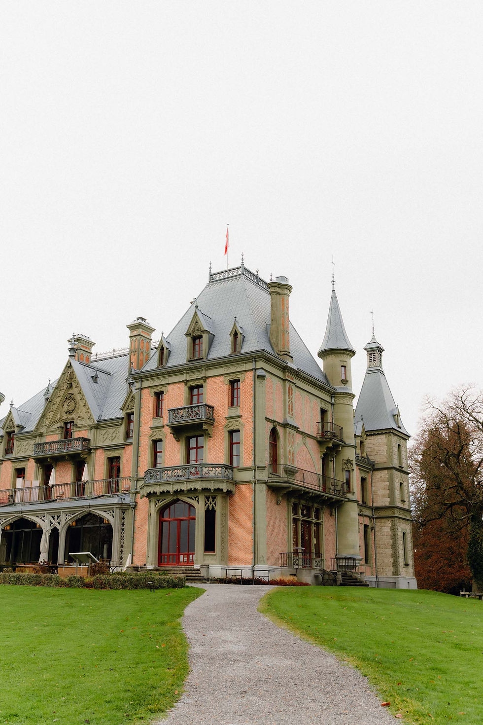 Großes, historisches Schloss mit Türmen und roter Fassade. Vorne ein Kiesweg, umgeben von grünem Rasen. Der Himmel ist bewölkt.