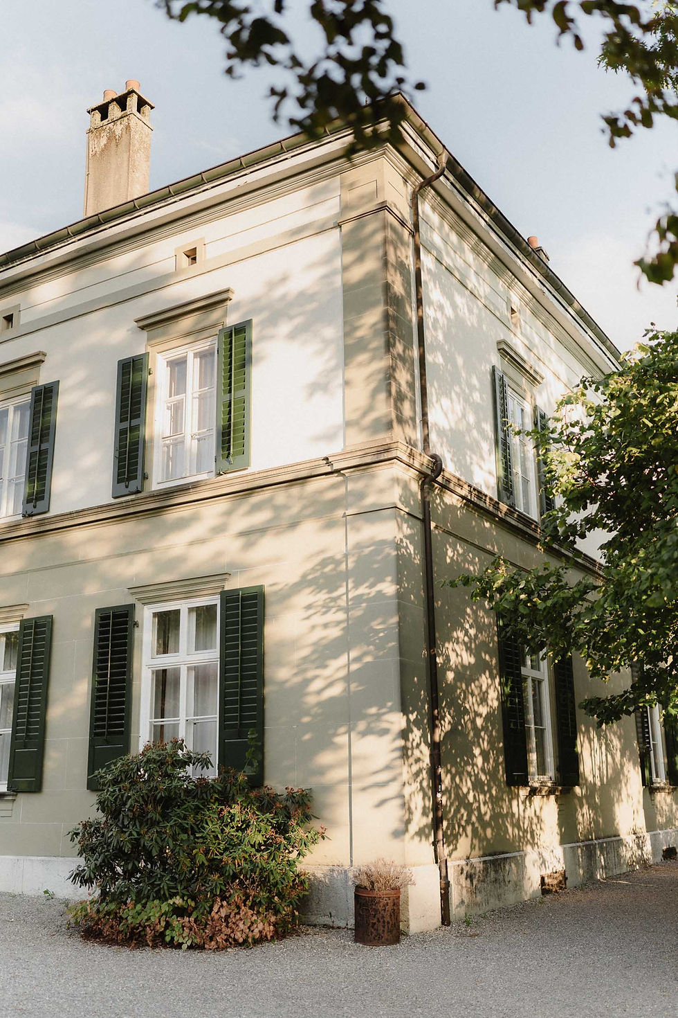 Sonniges Eckhaus mit hellen Fassaden, grünen Fensterläden und Baumschatten im Garten.