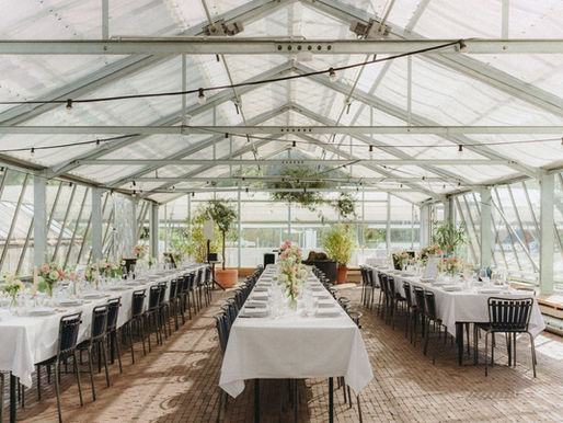 Langes Gewächshaus mit festlich gedeckten Tischen, weissen Tischdecken und Blumenarrangements. Helle, luftige Atmosphäre mit Pflanzen.