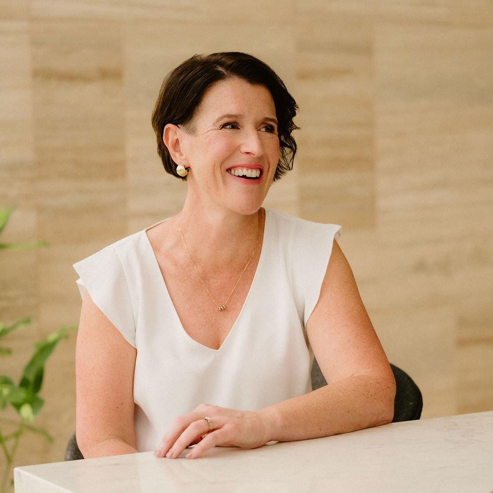 Sara looks off to the side, wearing a white sleeveless top with ruffled shoulders and a delicate necklace, seated at a table.