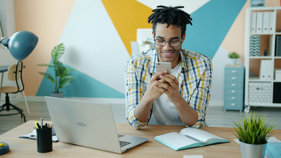 Man smiling at phone in colorful office, laptop open, notebook on desk, geometric wall art, vibrant and cheerful atmosphere.
