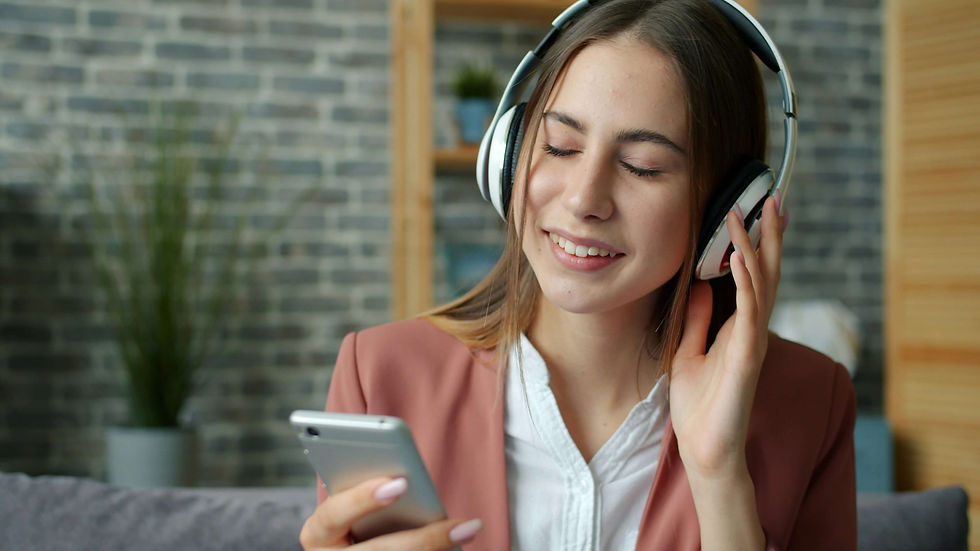Smiling woman in pink blazer listens to music with headphones, eyes closed and holding smartphone. Brick wall background with plants.