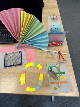 Models made of coloured paper sit on a the edge of a wooden school desk.