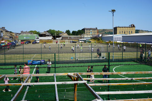 Children play soccer on a fenced green pitch beside an industrial lot, with a UNIQUE sign and clear blue sky.