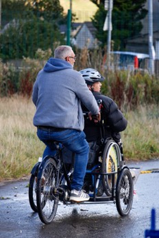 two people ride and adapted bicycle on a tarmaced area flanked to the left by a grassed area. One rider is pedaling the bike from the rear while the other sits in front in a wheelchair. 