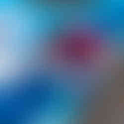 A person with blonde hair, wearing a pink jacket, white apron, blue gloves, and blue shoe covers is crouching down and painting a large outdoor mural. The mural features various shades of blue and white. The person is using a paint roller to apply light blue paint, with a paint tray beside them. The background shows more of the mural with abstract shapes in different blue tones.