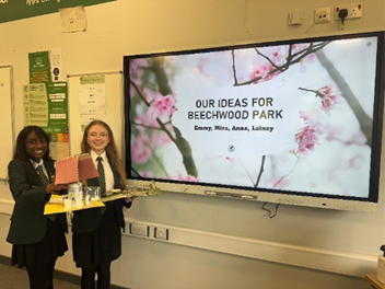 Two students in school uniforms stand in a classroom holding a model and papers. Behind them, a large screen displays the title 'OUR IDEAS FOR BEECHWOOD PARK' with the names Emmy, Mina, Anna, and Lainey, set against a background of pink cherry blossoms. Classroom posters and electrical outlets are visible on the wall.